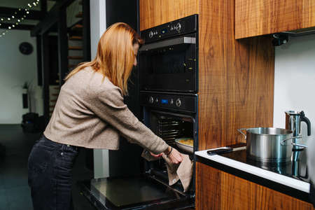Side view of a woman taking tray out of the oven using napkin to so she wouldn't get burned. In a big room with integrated kitchen.の写真素材
