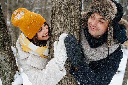 Happy couple in love look at each other's, hold on to a tree in the winter forest and laughing. They...
