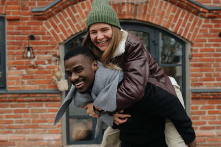 Carefree opposite sex interracial friends hanging out on the street. She is riding on his back. Both dressed warmly for late autumn. Both having a great time, smilingの写真素材
