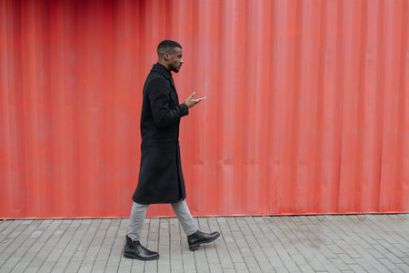 Side view of a sulky black man in a warm autumn coat walking in front of a red corrugated container...