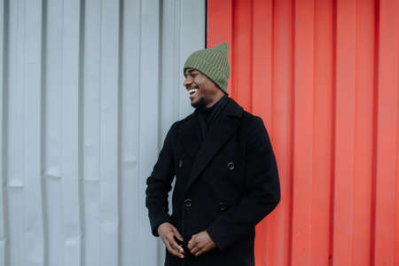 Laughing black man in a warm autumn coat standing in front of a red and white corrugated sheet container wall. His eyes closed.の写真素材