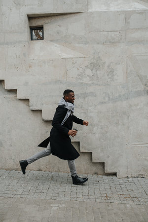 Uplifted black man in an autumn coat fast walking somewhere, holding a coffee cup in his hand. Over...
