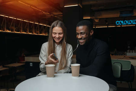 Happy smiling friends sitting in the cafe, sharing their one pair of headphones. They have different sex and race.の写真素材