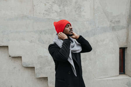 Fancy black man in a warm autumn coat putting on a wibrant red knitted hat. He is standing in front of the wall, waiting for someone.の写真素材