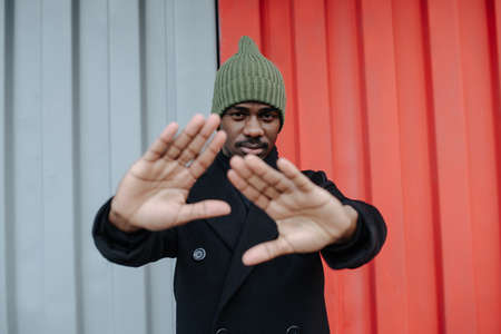 Insistend black man in a warm autumn coat blocking camera with his hands. He is standing in front of a red and white corrugated sheet container wall.の写真素材