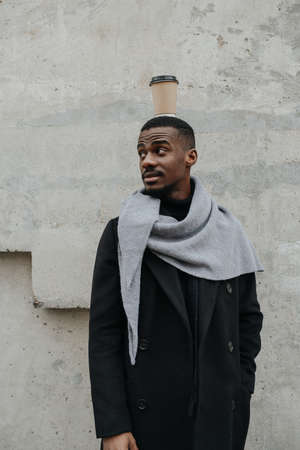 Silly funny black man in a coat standing imperturbable with a coffee cup on his head. He is waiting for someone.の写真素材