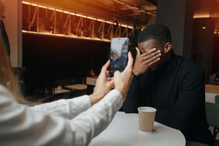 Woman taking a picture of her shy friend in a cafe, he is covering his face with a hand. They have different sex and race.の写真素材