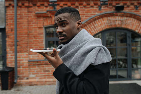 Talking on a loud speaker black man standing on the street at autumn. He is warmly dressed in a coat and a scarf. Red bricked building in background.の写真素材