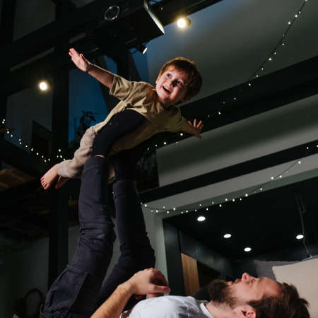 Square image of father lifting his happy child with his legs. He is spreding his arms like a plane. Upward angle. Garlands under the ceiling.の写真素材