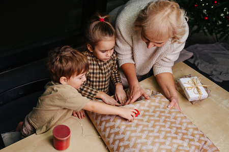 Methodical grandmother teaches granddaughter how to wrap a present, while little boy plays with a...