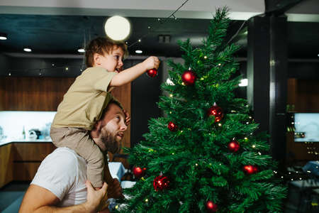 Adorable toddler son sitting on her dad's shoulders decorating a christmas tree with red shiny balls. Dad looking up at what she is doing.の写真素材