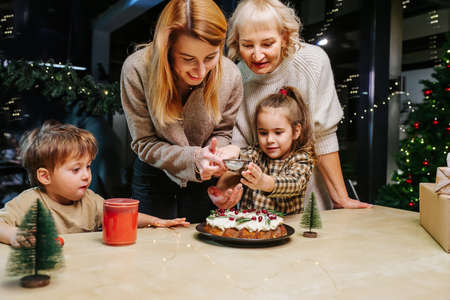 Overjoyed family preparing a cake for the christmas. Sprinkling it with sugar powder. Lit with a yellow lamp. Dark decorated christmas tree in background.の写真素材