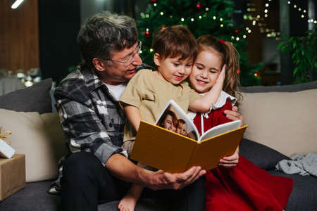 Grandpa sitting on a sofa looking through a photo album with his grandchildren. Two little siblings enjoy pictures. Christmas tree in background.の写真素材