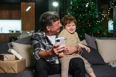 Fun grandpa sitting on a sofa, trying to take selfie with his giggling toddler grandson sitting on his knees. Christmas tree in background.の写真素材