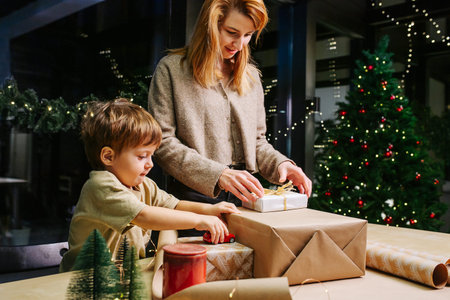 Mother prepares for christmas, wrapping presents in paper. Her son is keeping her company, playing...