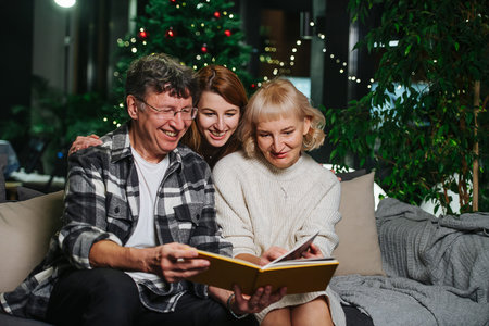 Elder parents and their adult daughter sitting on a sofa looking through a photo album. Christmas...