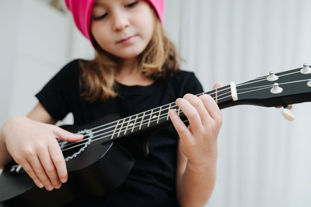 Focused pretty little girl in a pink winter hat playing on a guitalele at home. Focus on her fingers.の写真素材