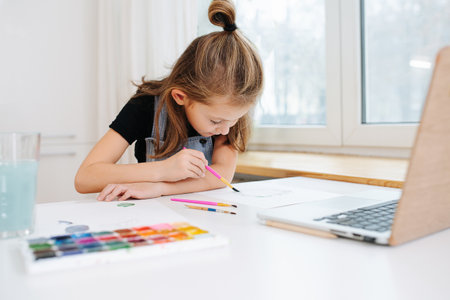 Focused immersed little girl sitting behing a big table next to a window, painting with watercolor in front of a laptop, using it as a template. low angle.の写真素材