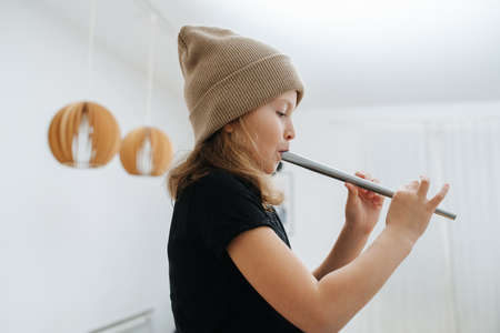 Side view of a little girl in a beige hat learning to play on an Irish flute. At home in a mostly white room.の写真素材