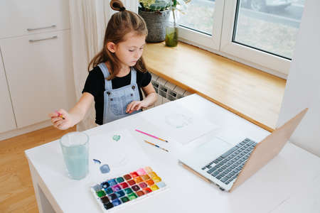 Serious looking little girl sitting behing a big table next to a window, painting with watercolor in front of a laptop, using it as a template. High angle.の写真素材