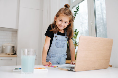 Smiling little girl leaning on a table, posing for a photo, looking in the camera. She is painting...