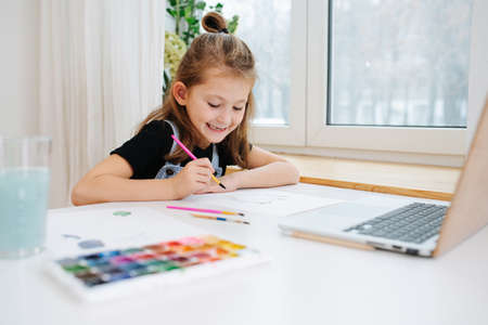 Immersed happy little girl sitting behing a big table next to a window, painting with watercolor in front of a laptop, using it as a template. low angle.の写真素材