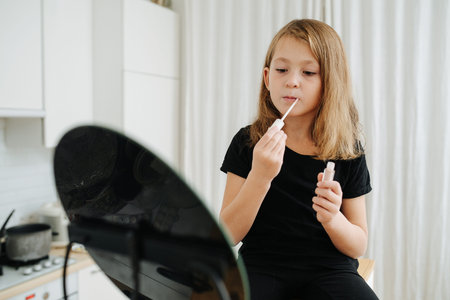 Serious little girl sitting in the kitchen, looking at the mirror, applying lipstick with a brushの写真素材