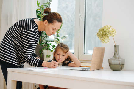 Mom helping little girl to draw. She's standing over the table, drawing with pen. Girl is watching carefully, leaning on her hand.の写真素材