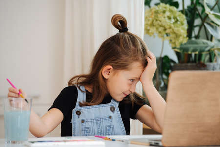 Playful little girl sitting behing a big table next to a window, painting in front of a laptop, using it as a template. She is showing of, putting on an act in theatrical fashion.の写真素材