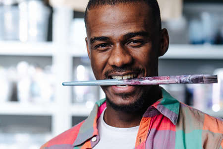 Portrait of a silly black man in colorful shirt biting his teeth on a brush in a workshop. All kinds of containers on shelves in backgound.の写真素材