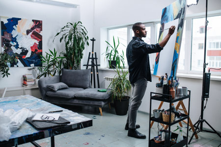 Serious black man painting on a blank sheet on an easel inside of his workshop room in the apartment. A sofa and an abstract painting on the wall behind. Window view on the building next door.の写真素材