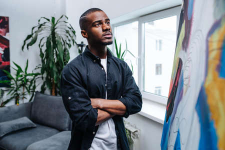 Impressionable black man looking at an easel with his unfinished painting inside of his apartment.の写真素材