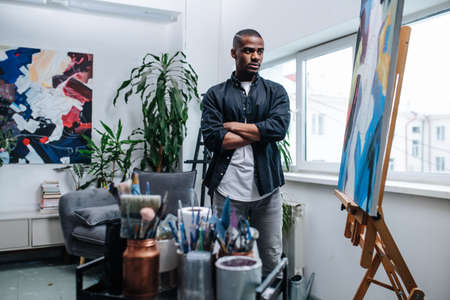 Critical black man looking at an easel with his unfinished painting inside of his workshop in apartment. A sofa and an abstract painting on the wall behind. Window view on the building next door.の写真素材