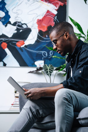 Side view african american man sitting on a sofa with a laptop in his apartment. An abstract painting behind him.の写真素材