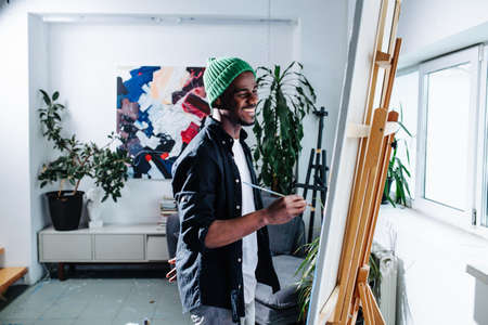 A happy black man painting on an easel inside of his apartment. Lots of potted plants and an abstract painting on the wall.の写真素材