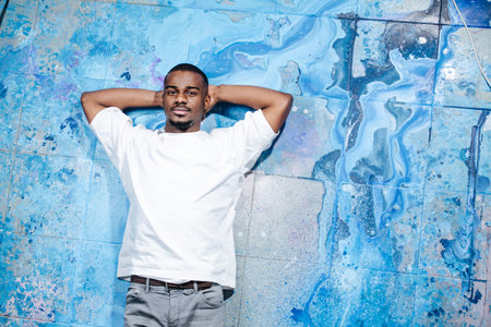 Looking at the camera black male artist lying on the blue paint stained floor. With hands behind his...