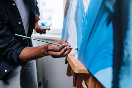 Close up view black man painting on an easel inside of his apartment. He is coloring it. Window view on the building next door.の写真素材