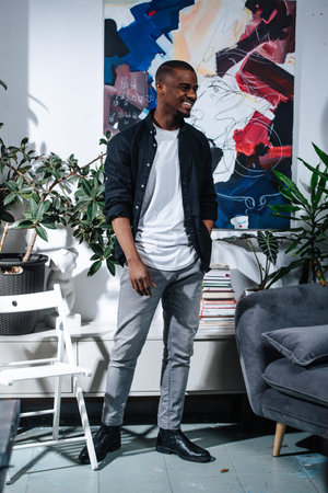 Cheerful african american painter standing in front of his abstract painting hanging on the wall. Potted plants and furniture around him.の写真素材