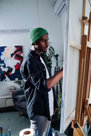 Focused peaceful black man painting on an easel inside of his messy disorderly apartment. An abstract painting on the wall behind.の写真素材