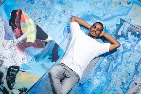 Black male artist lying on blue paint stained floor next to an abstract painting. Looking at the...