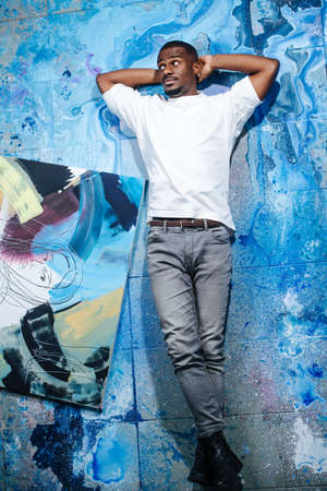 Looking at the side chill black male artist lying on blue paint stained floor next to an abstract painting. Leg on leg, hands behind his head.の写真素材