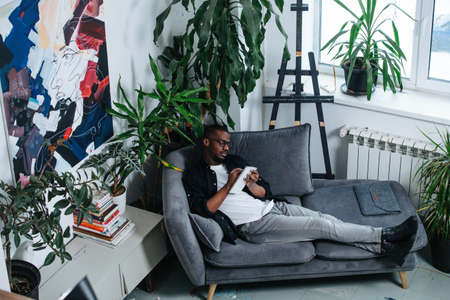 African american man chilling on a sofa in the corner of his apartment. He is writing notes, surrounded by potted plants.の写真素材