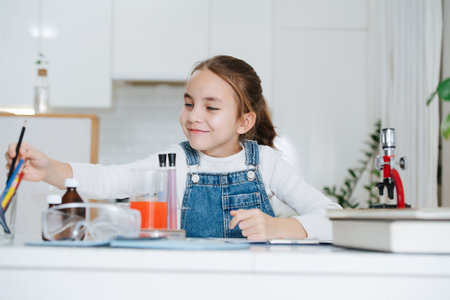 Smiling little girl doing home science project, reaching for crayons. She has chemistry glassware with colorful liquids and microscopeの写真素材