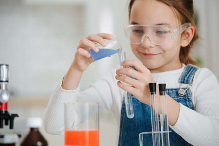Smiling confident girl doing home science project, pouring liquid into a flask. She is wearing plastic glasses and has glass chemical equipment and microscopeの写真素材
