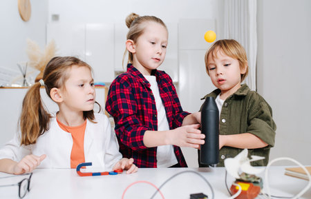 Three kids doing home science project, hovering  ball with a fan. All standing behind the table,...