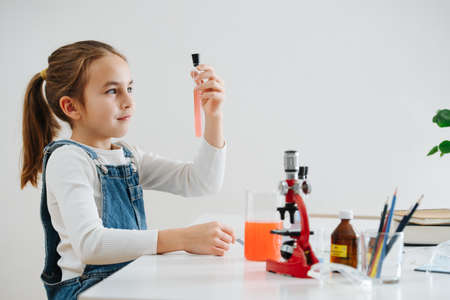 Mermerized girl doing home science project, looking at pale red liquid in the flask. She has glass chemical wares equipment and microscopeの写真素材