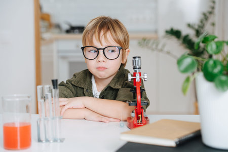 Uneasy little boy in glasses sitting behind the table in front microscope