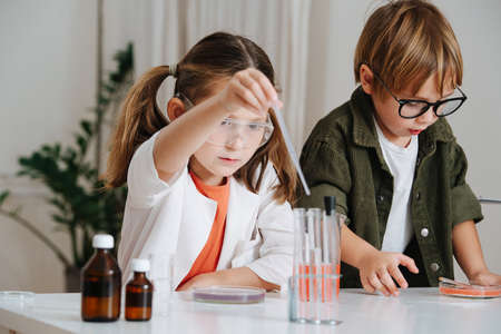 Focused little girl in white robe filling a pipette next to her boy picks a dishの写真素材