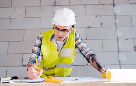 Focused hard working construction engineer in white helmet drawing on a blueprint on an improvised board table. Over a rough gray brick wall.の写真素材