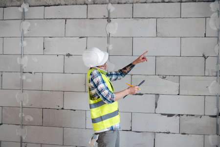 Busy construction engineer in a white helmet discussing problems on the phone, pointing at a brick wall, holding a clipboard.の写真素材